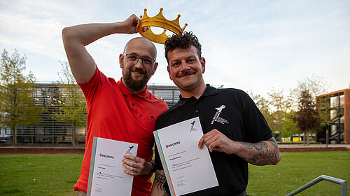 Die beiden stehen nebeinander. Sie halten in der linken Hand ihre Urkunden. Luca hält eine goldene Krone aus Plastik über beiden Köpfen.