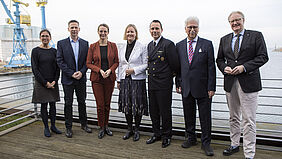 Die sieben Personen stehen nebeneinander. Im Hintergrund ist das Wasser der Wismarbucht und zwei Hafenkäne zu sehen.
