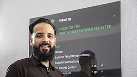 Mann mit Bart und dunklem Poloshirt steht vor schwarzem Bildschirm mit grüner und weißer Schrift im Fakultätsgebäude auf dem Campus