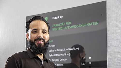 Mann mit Bart und dunklem Poloshirt steht vor schwarzem Bildschirm mit grüner und weißer Schrift im Fakultätsgebäude auf dem Campus