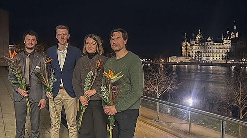 Die vier Personen stehen im Dunkeln auf einer Terasse. Im Hintergrund ist das angestrahlte Schweriner Schloss zu sehen.