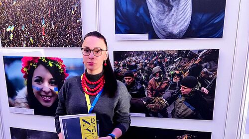 Dr. Anna Fomenko, mit roter Halskette und einem Programmheft der Cafe Kyiv Veranstaltung steht vor einer Bilderwand, die Eindrücke aus der neuen Lebensrealität ukrainischer Staatsbürger widerspiegeln. Zwischen Hoffnung und Leid.