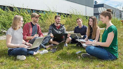 Im Bild sind sechs Studierende der Wirtschaftsinformatik zu sehen, die mit ihren Laptops auf der Campuswiese vor dem Haus 6 sitzen.