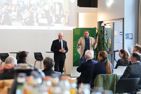 Two men are standing in front of an audience in a modern event space. One is wearing a black suit and holding a glass, while the other is wearing a light-coloured blazer and a blue scarf and is speaking animatedly. A screen showing a video conference can be seen in the background.