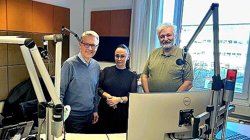 Three people are standing in a radio studio with microphones and a mixing desk, presenting the EUComplianceM4UA project on Radio LOHRO.