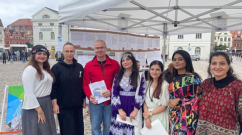 Frank Junge steht mit Dr. Anna Fomenko und fünf Studentinnen unter einem Pavillon auf dem Wismarer Marktplatz.