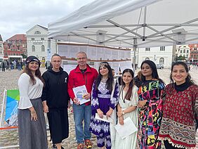 Besuch am Hochschulstand Frank Junge steht mit Dr. Anna Fomenko und fünf Studentinnen unter einem Pavillon auf dem Wismarer Marktplatz.
