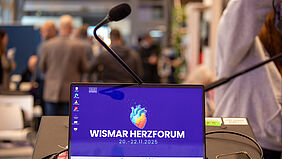 Auf dem Bildschirm eines Laptops ist eine Herzgrafik zu sehen. Dahinter im Raum sind unscharf Menschen im Gespräch zu erkennen.