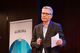Ein Redner steht auf einer Bühne mit Headset-Mikrofon und präsentiert vor einem Aurora-Banner beim Aurora Symposium on Global Citizenship 2026.