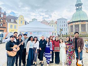 Internationale Studierende auf dem Marktplatz. Eine Gruppe Studierender steht mit Musikinstrumenten vor einem Pavillon auf dem Wismarer Marktplatz.