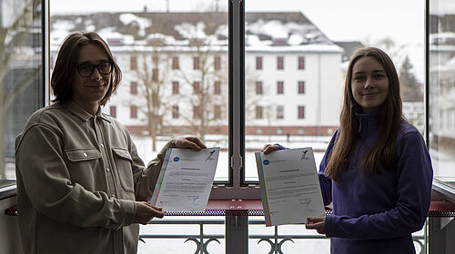 Die beiden Studierenden stehen vor einem offenen Fenster und halten ihre Urkunden in den Händen. Im Hintergrund ist Haus 19 zu sehen, in dem sie studieren.
