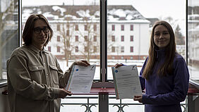 Die beiden Studierenden stehen vor einem offenen Fenster und halten ihre Urkunden in den Händen. Im Hintergrund ist Haus 19 zu sehen, in dem sie studieren.