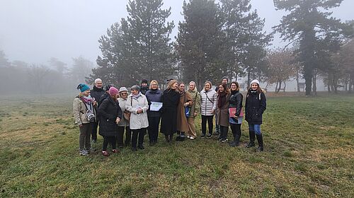 Projektpartner:innen bei der Erkundung des Pilotstandorts in Érd Gruppe von rund 18 Personen steht in herbstlicher Kleidung auf einer leicht nebligen Wiese vor einer Baumreihe. Einige halten Mappen oder Unterlagen in der Hand.