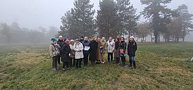 Projektpartner:innen bei der Erkundung des Pilotstandorts in Érd Gruppe von rund 18 Personen steht in herbstlicher Kleidung auf einer leicht nebligen Wiese vor einer Baumreihe. Einige halten Mappen oder Unterlagen in der Hand.