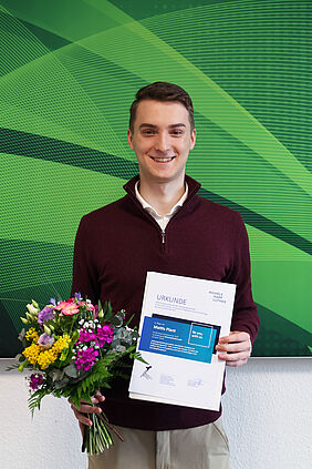Mattis Plant hält Urkunde und Blumenstrauß nach der Auszeichnung seiner Bachelorarbeit.