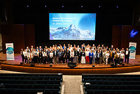 Rund 100 internationale Teilnehmende stehen für ein Gruppenfoto auf einer Bühne vor einer großen Präsentationsleinwand des Aurora Symposiums.