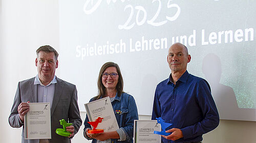 Die drei Personen sthen nebeneinander und halten ihre Urkunde sowie je einen Fischerpokal in Fakultätsfarbe in den Händen. Hinter Ihnen ist auf einem Bildschirm der Spruch "Spielerisch Lehren und Lernen" zu sehen.