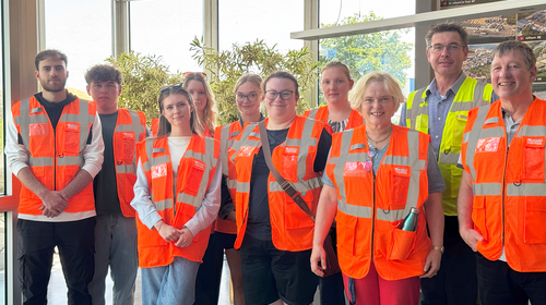 Group photo of students wearing orange high-visibility vests in the entrance hall. The individuals are standing in a line. Mr Mertens, the contact person for the company visit, is second from the right.