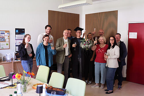Dominic Strube steht nach seiner erfolgreichen Promotion im Kreis von Kolleginnen und Kollegen. Die Gruppe hält Sektgläser in den Händen und feiert den Abschluss der Dissertation. Dominic Strube trägt einen Doktorhut und hält einen Blumenstrauß.