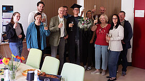 Erfolgreiche Promotion von Dominic Strube gefeiert Dominic Strube steht nach seiner erfolgreichen Promotion im Kreis von Kolleginnen und Kollegen. Die Gruppe hält Sektgläser in den Händen und feiert den Abschluss der Dissertation. Dominic Strube trägt einen Doktorhut und hält einen Blumenstrauß.
