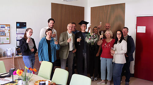 Celebration of Dominic Strube’s successful PhD. Dominic Strube stands amongst his colleagues following the successful completion of his PhD. The group is holding champagne glasses and celebrating the completion of his thesis. Dominic Strube is wearing a doctoral cap and holding a bouquet of flowers.