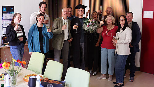 Dominic Strube stands amongst his colleagues following the successful completion of his PhD. The group is holding champagne glasses and celebrating the completion of his thesis. Dominic Strube is wearing a doctoral cap and holding a bouquet of flowers.