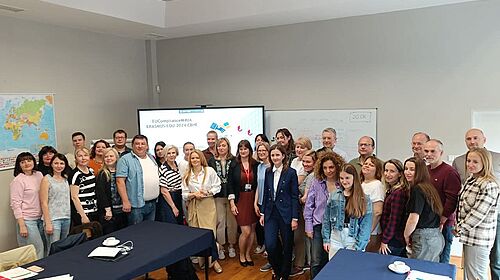 Etwa 40 Personen aus verschiedenen Ländern stehen gemeinsam in einem Seminarraum vor einem Bildschirm mit dem Titel des EU-Projekts EUComplianceM4UA.
