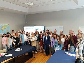 Etwa 40 Personen aus verschiedenen Ländern stehen gemeinsam in einem Seminarraum vor einem Bildschirm mit dem Titel des EU-Projekts EUComplianceM4UA.