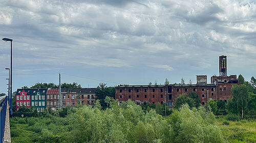 barrierefrei: Es ist ein verfallenes mehrstöckiges Backsteingebäude mit teilweise zerstörten Fenstern und angrenzenden bunten Wohnhäusern zu sehen - vor bewölktem Himmel und grüner Vegetation im Vordergrund.