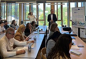 Teilnehmer:innen eines Projektmeetings sitzen an Tischen in einem hellen Seminarraum mit Blick nach draußen, während vorne ein Vortragender spricht.