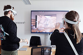 Zwei Studierende mit Apple Vision Pro Brillen stehen in einem Seminarraum. Eine große Leinwand zeigt virtuelle Inhalte. Eine Person hebt beide Hände, um mit der VR-Umgebung zu interagieren.