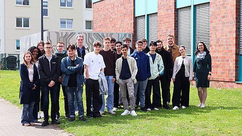 Gruppe von Schülerinnen und Schülern steht gemeinsam mit Lehrenden vor einem Hochschulgebäude auf einer Grünfläche.