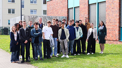 Gruppe von Schülerinnen und Schülern steht gemeinsam mit Lehrenden vor einem Hochschulgebäude auf einer Grünfläche.
