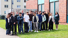 Gruppe von Schülerinnen und Schülern steht gemeinsam mit Lehrenden vor einem Hochschulgebäude auf einer Grünfläche.