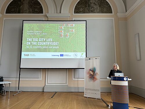 Eine Frau spricht an einem Rednerpult vor einem großen Bildschirm mit der Konferenzfolie „The Big City Life on the Countryside? Yes, AI, creativity, nature and people“ in einem historischen Saal.