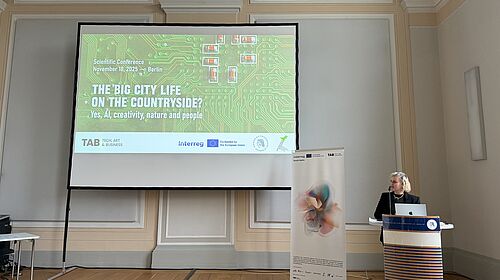 Conference kick-off at the Humboldt Graduate School. A woman speaks at a lectern in front of a large screen displaying the conference slide ‘The Big City Life on the Countryside? Yes, AI, creativity, nature and people’ in a historic hall.