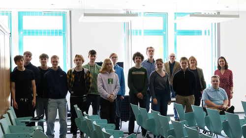 Group photo of around 17 people including pupils, teachers, and university staff in a seminar room with turquoise chairs. The group stands and sits in two rows in front of large windows.