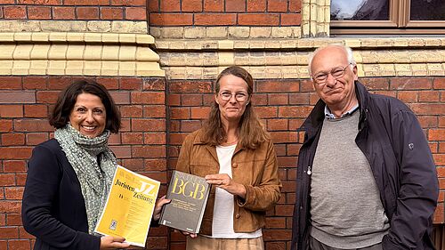 Three people are standing in front of a brick façade; two of them are holding specialist legal publications.