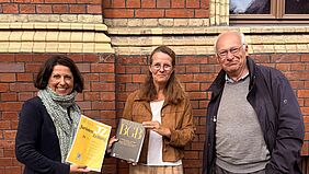 Forschung zum Umwelthaftungsrecht vorgestellt Drei Personen stehen vor einer Backsteinfassade; zwei von ihnen halten juristische Fachpublikationen in den Händen.