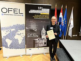 PhD Laima Gerlitz with certificate of attendance of the 11th OEFL conference in front of an event poster in the meeting room.