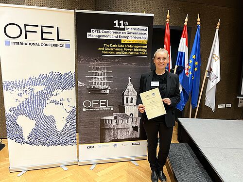 PhD Laima Gerlitz with certificate of attendance of the 11th OEFL conference in front of an event poster in the meeting room.