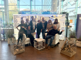 Zehn Personen, darunter Männer und Frauen, stehen und sitzen in zwei Reihen vor einem Banner mit der Aufschrift „Gdynia Miasto Modernizmu“. Im Hintergrund: große Fensterfront mit Blick auf das Stadtzentrum von Gdynia.
