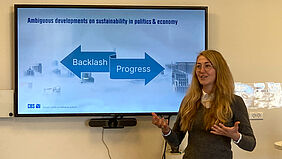 Johanna steht mit gestikulierenden Händen direkt vor einem Bildschirm. Auf dem ist eine grafische Struktur, die mit englischen Begriffen gefüllt ist, zu sehen.