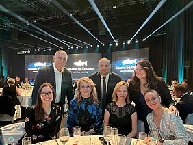 Netzwerkstärkung beim „Salmon Evening“ Eine Gruppe von sieben Personen sitzt und steht an einem festlich gedeckten Tisch in einer Veranstaltungshalle mit Bühne im Hintergrund.