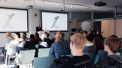 Wir schauen über die Köpfe der Schüler_innen hinweg, die in einem Raum sitzen und nach vorn auf zwei Bildschirme und den Redner schauen.