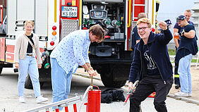 Studierende probieren vor einem Feuerwehrfahrzeug eine Handdruckspritze aus.