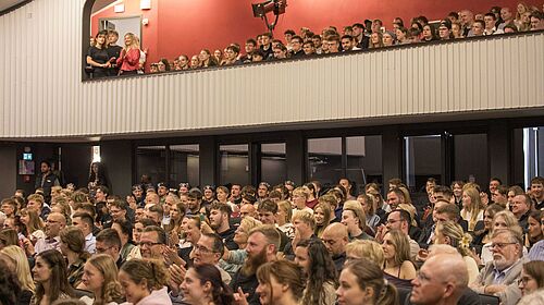 Publikum in einem Theater bei einem Festakt, sitzend und stehend auf zwei Ebenen. Sie blicken aufmerksam nach vornund manche klatschen.