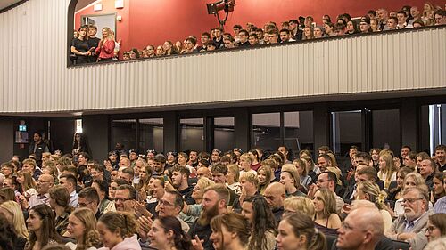 Publikum in einem Theater bei einem Festakt, sitzend und stehend auf zwei Ebenen. Sie blicken aufmerksam nach vornund manche klatschen.