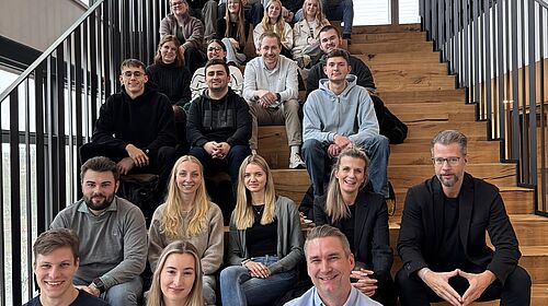 About 25 people are sitting on a wide wooden staircase in a light-filled room, smiling at the camera.
