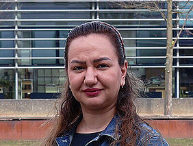 Porträt einer Frau im Freien vor einem modernen Hochschulgebäude. Sie trägt eine Jeansjacke und blickt in die Kamera.